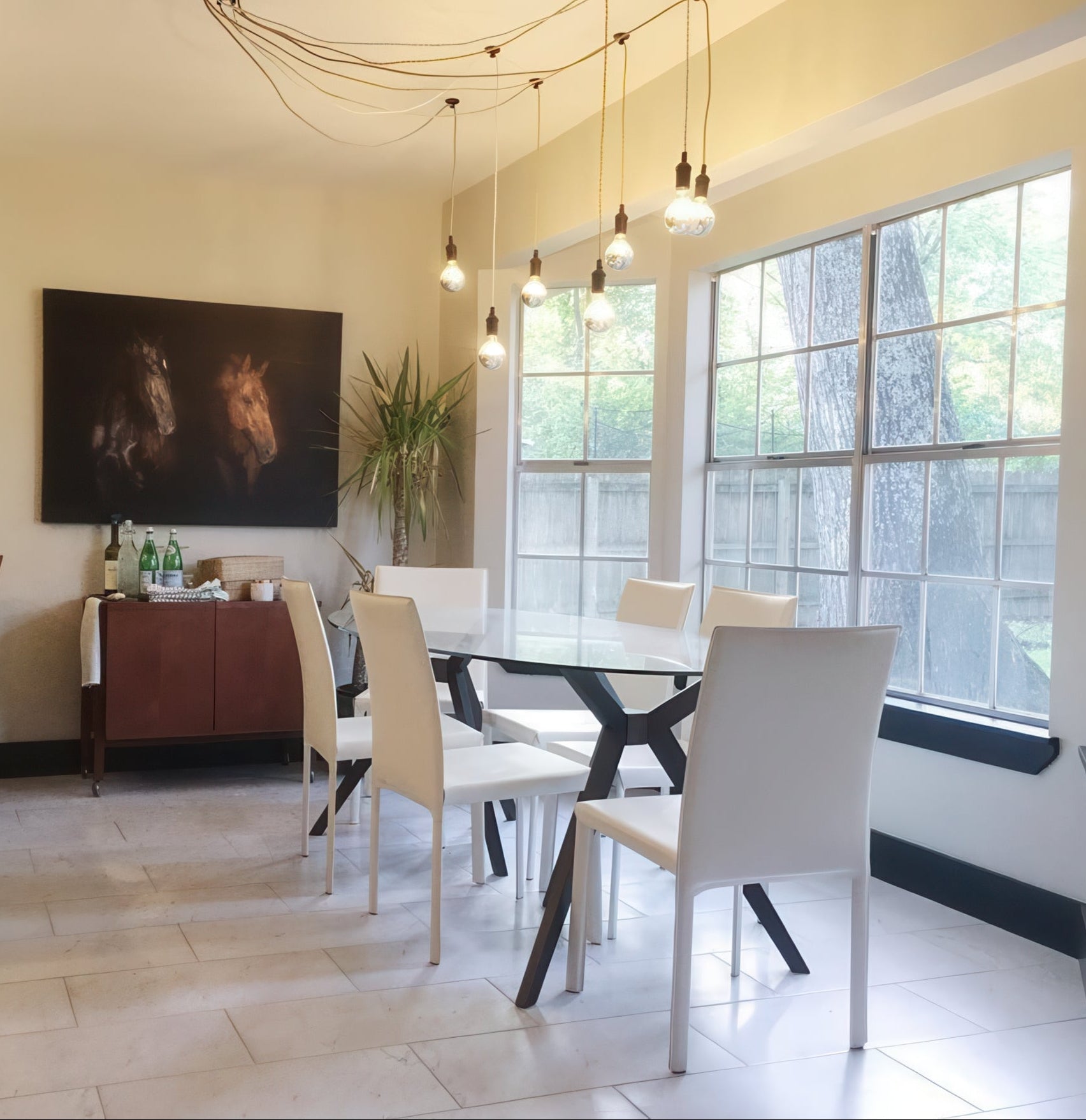 Modern dining room with a glass table and white chairs, featuring a large window and artwork on the wall. off center ceiling light swag over dining table