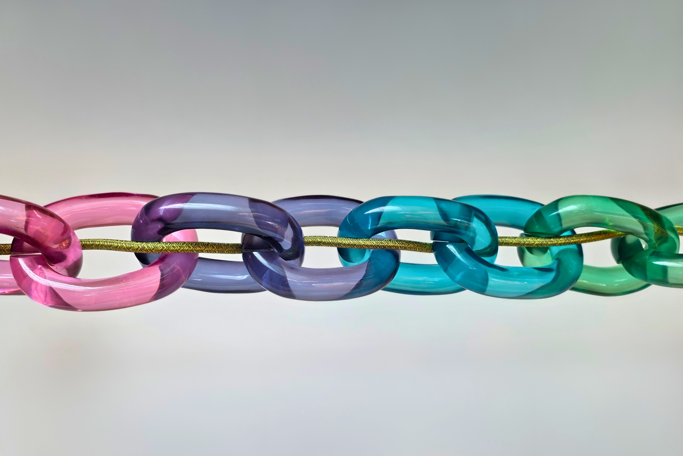 A close-up of a translucent acrylic chain link garland hanging vertically. The links transition through pink, purple, blue, teal, and green shades, suspended on a thin gold cord against a soft, light beige background.