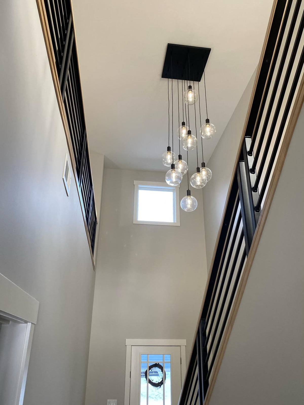 A modern multi-light chandelier with clear glass globe bulbs and a black rectangular ceiling canopy, installed in a high-ceilinged staircase hall with white walls and black-and-wood railings.
