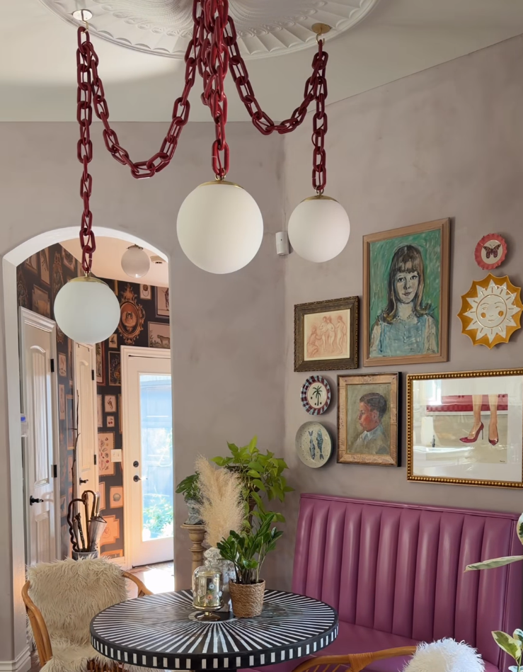 A dining area featuring a unique red chain chandelier with three white spherical glass globes hanging from a decorative white ceiling. The room has a pink tufted bench, a black-and-white patterned table, and a gallery wall filled with framed art.