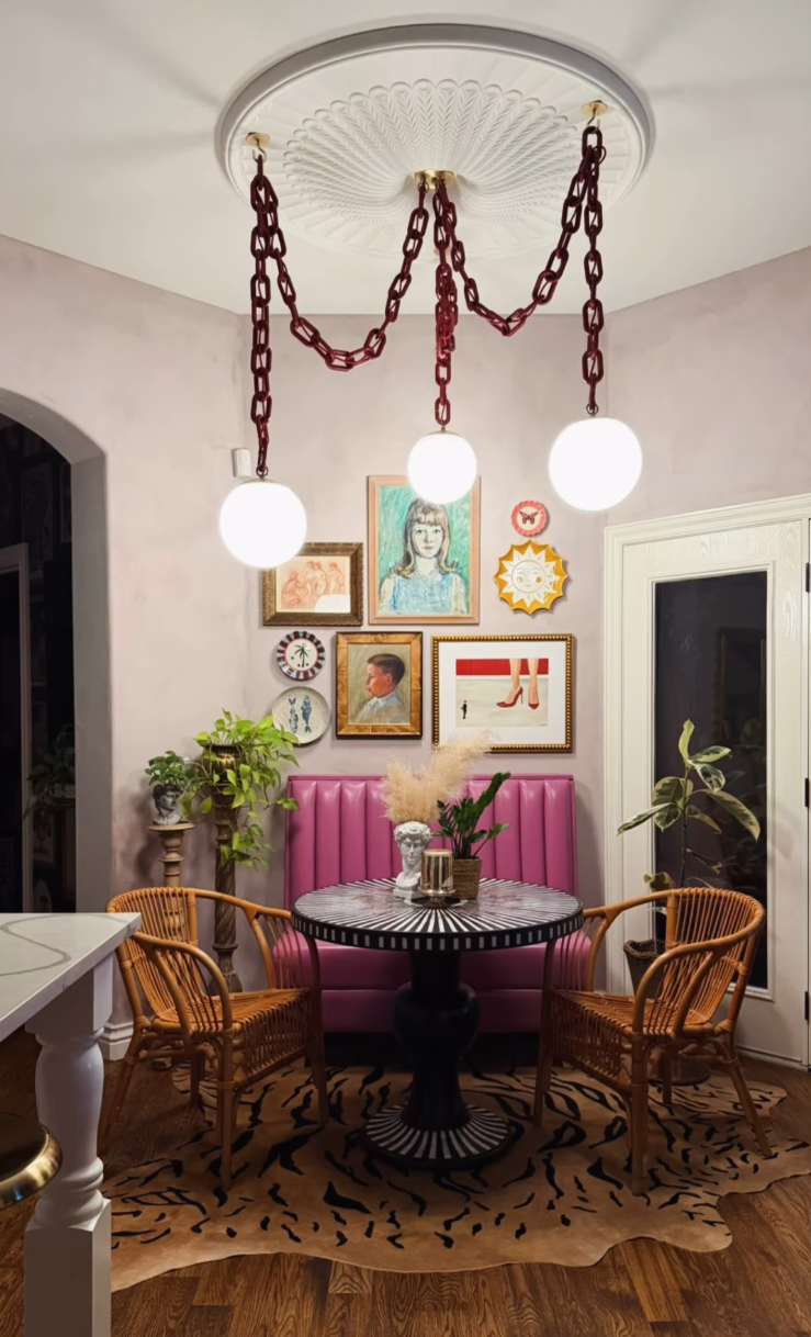 A bohemian-style dining area featuring a unique three-light pendant chandelier with dark red chain links and white globe shades, hanging from a white decorative ceiling medallion. The room has pink walls, a pink tufted banquette, two wicker chairs, and a patterned rug.