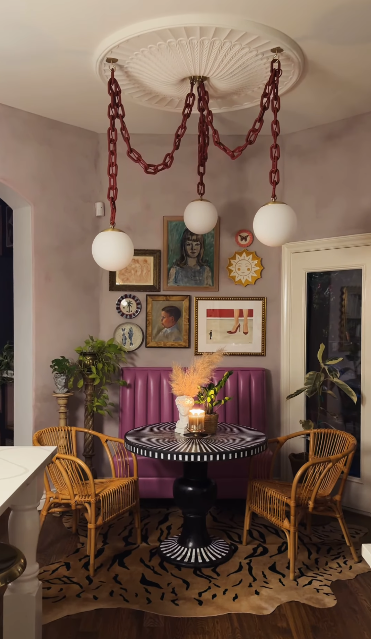 A styled dining nook featuring a round black-and-white patterned table, two wicker chairs, a purple tufted bench, and a decorative ceiling fixture with a white ornate medallion, red chains, and three white globe shades.